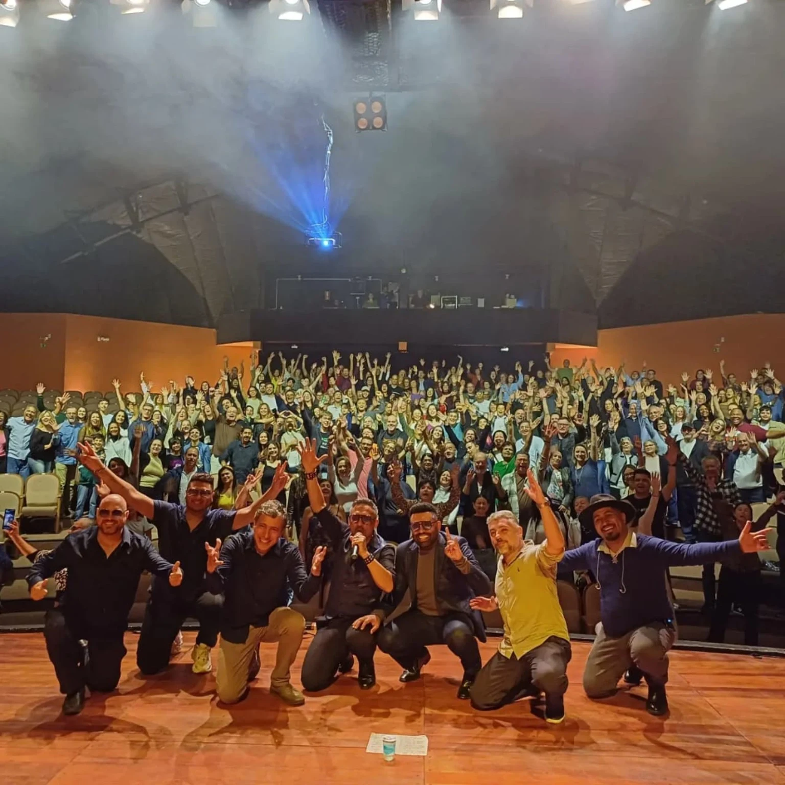 Banda no palco de um teatro com o público ao fundo, de braços levantados